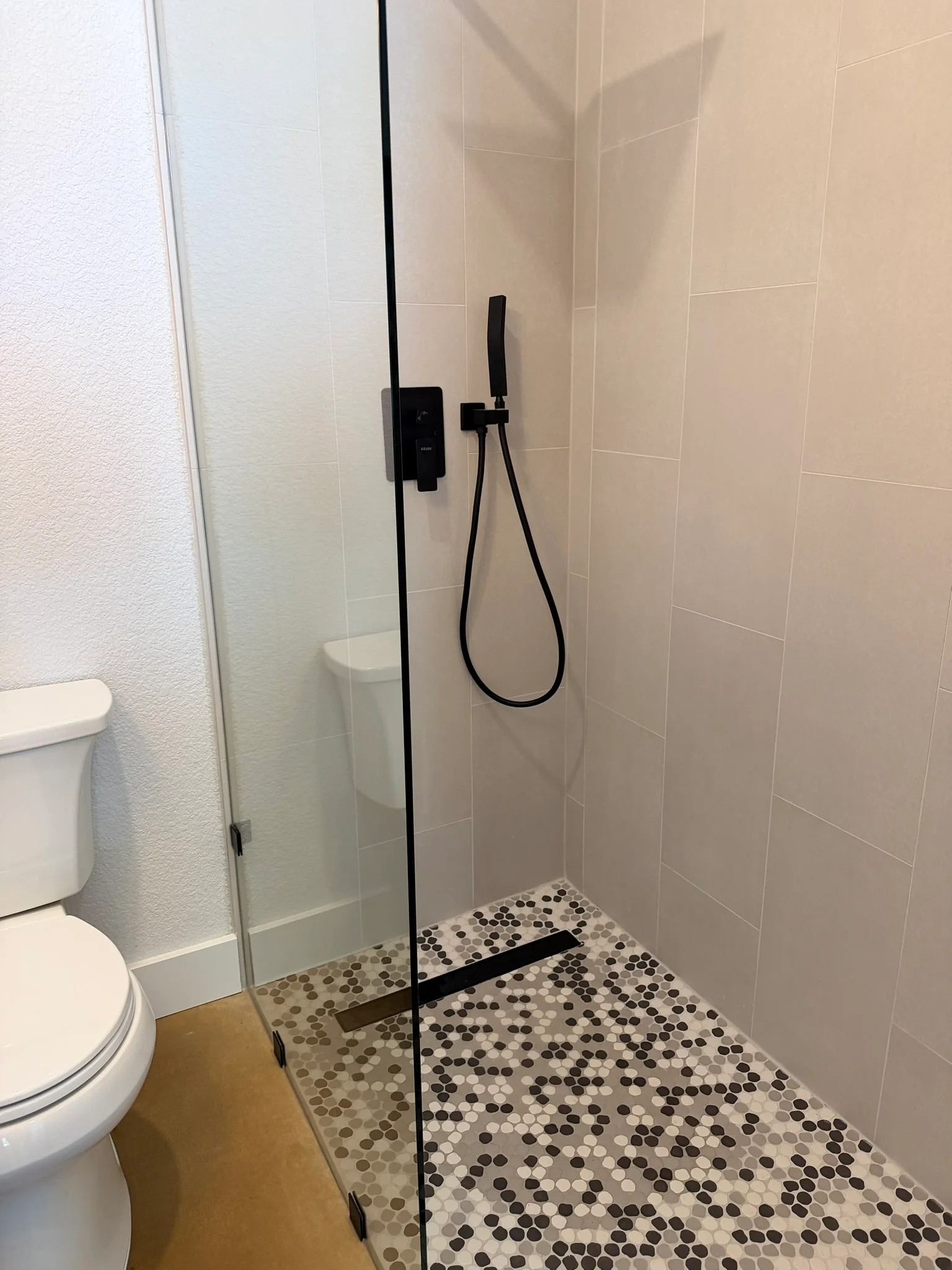 Walk-in shower with beige tile walls, penny tile mosaic floor, frameless glass door, and black fixtures